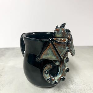 May include: A black ceramic mug with a dragon design. The dragon has brown and teal wings and a tail that wraps around the mug. The mug has a handle and is on a white background.