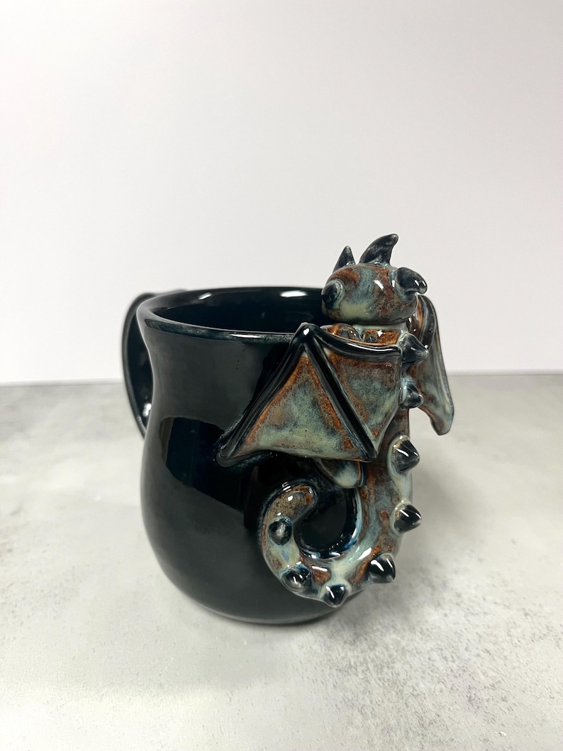 May include: A black ceramic mug with a dragon design. The dragon has brown and teal wings and a tail that wraps around the mug. The mug has a handle and is on a white background.