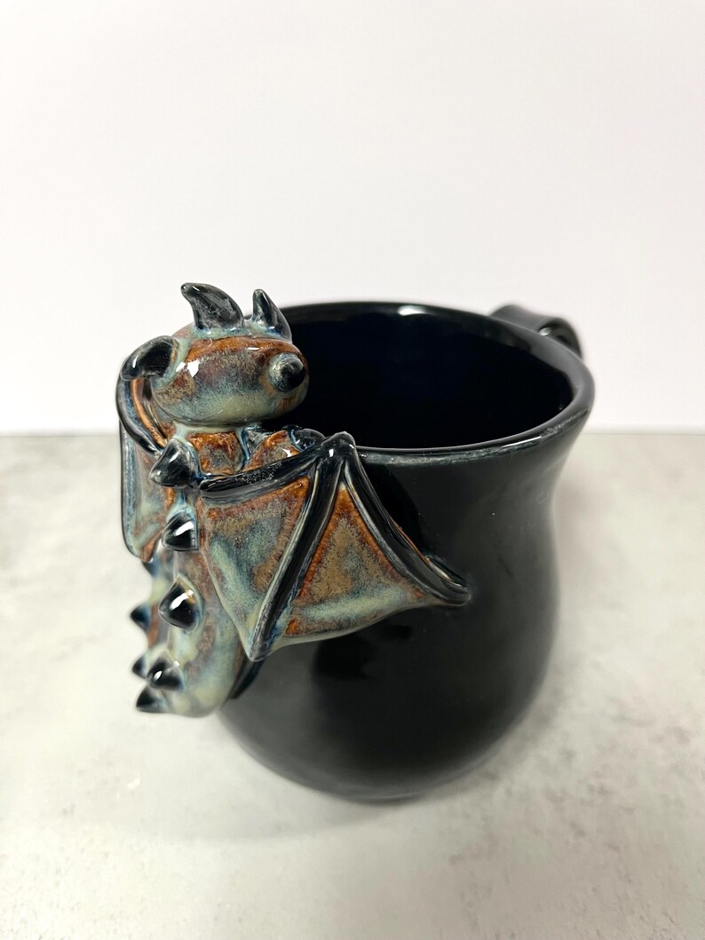 May include: Black ceramic mug with a bat-shaped handle. The bat has brown, blue, and tan accents. The mug is glossy and has a dark interior. The mug is a unique, handcrafted item.