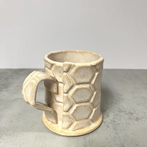 May include: A white ceramic mug with a hexagonal pattern. The mug has a handle and a rounded base.