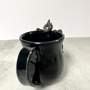May include: Black ceramic mug with a glossy finish. A small, detailed dragon figure is perched on the rim. The dragon has brown, blue, and green accents. The mug has a large handle.