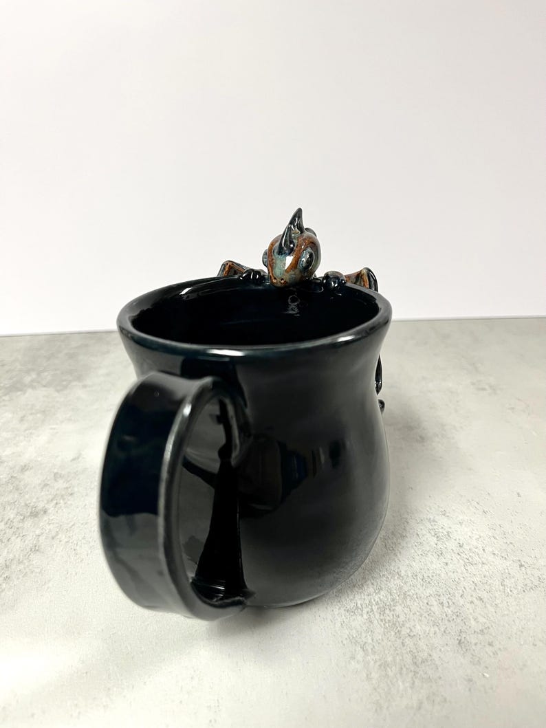 May include: Black ceramic mug with a glossy finish. A small, detailed dragon figure is perched on the rim. The dragon has brown, blue, and green accents. The mug has a large handle.