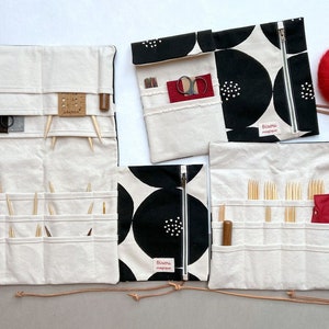 以下が含まれることがあります： クリーム色と黒色の生地で作られた編み針ケースのコレクション。白と黒の円形模様が特徴です。ケースは開いており、編み針、ハサミ、その他のツールが見えます。赤い毛糸の玉も2つあります。