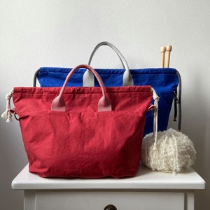 Pode incluir: Duas sacolas de lona, uma vermelha e outra azul, com um novelo de lã branca e agulhas de tricô em uma mesa branca.