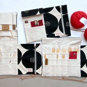 May include: A collection of cream-colored canvas organizers for knitting needles and accessories, featuring black and white circular patterns, zippered pouches, and two round balls of bright red yarn with white labels, all arranged on a flat surface.