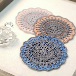 Puede incluir: Tres doilies de ganchillo en tonos azules y rosas, con una figurita de cisne de cristal en primer plano.