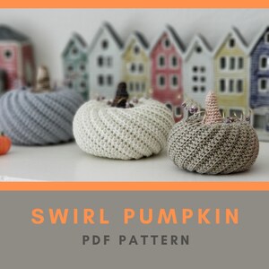 May include: Three crocheted swirl pumpkins in gray, white, and brown. The pumpkins are sitting on a white surface in front of a row of miniature houses. The text "SWIRL PUMPKIN PDF PATTERN" is displayed below the pumpkins.