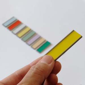 May include: A rectangular yellow plastic tag with a thin gray border held in the foreground, with a series of seven additional rectangular tags in various colors including light blue, orange, white, yellow, green, lavender, and beige arranged behind.