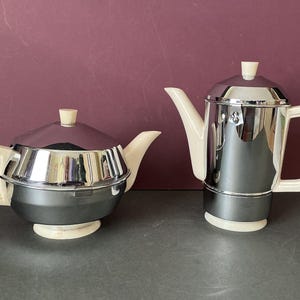 May include: Two vintage teapots with chrome and cream-coloured accents. One is a rounded shape with a lid and handle, the other is a tall, rectangular shape with a spout and handle. Both have a retro design.