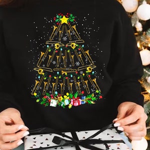 Op de afbeelding: Een zwart sweatshirt met een kerstboomontwerp gemaakt van gouden trombones. De boom is versierd met rode en groene kerstballen en witte lampjes.