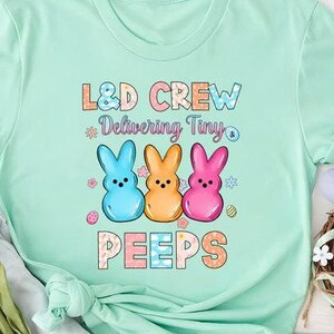 Może przedstawiać: Jasnozielony t-shirt z tekstem "L&D Crew Delivering Tiny Peeps" w kolorowej czcionce. Projekt zawiera trzy kreskówkowe pisklęta w różnych kolorach.