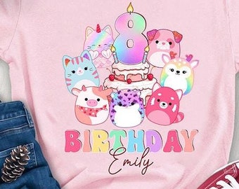 以下が含まれることがあります： ピンクのTシャツに、カラフルなグラフィックがプリントされています。8本のろうそくが立てられた誕生日ケーキと、たくさんの漫画の動物が描かれています。グラフィックの下には「Birthday Emily」という文字がプリントされています。