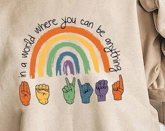 May include: A beige sweatshirt with a rainbow and the text "in a world where you can be anything" printed on the front. The rainbow is made up of different coloured stripes and has a purple, blue, and green inner rainbow. Below the rainbow are five hands in different hand signs.