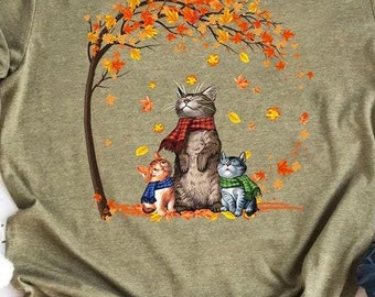 Könnte beinhalten: Ein grünes T-Shirt mit einem Motiv von drei Katzen, die unter einem Baum mit orangefarbenen und gelben Blättern stehen. Die Katzen tragen Schals und die größte Katze schaut nach oben.