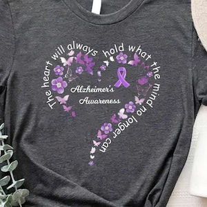 May include: A dark gray t-shirt with a purple ribbon and butterflies. The text on the shirt reads "The heart will always hold what the mind no longer can. Alzheimer's Awareness."