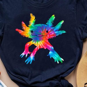 Könnte beinhalten: Ein marineblaues T-Shirt mit einem farbenfrohen Tie-Dye-Axolotl-Design. Der Axolotl ist in leuchtenden Regenbogenfarben dargestellt, mit einem lächelnden Gesicht und detaillierten Merkmalen. Das T-Shirt hat kurze Ärmel und einen Rundhalsausschnitt.