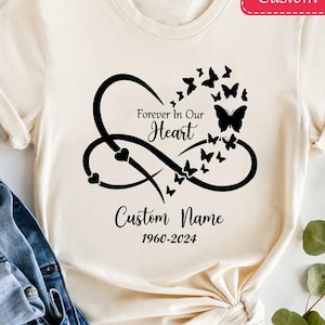 May include: A white t-shirt with a black infinity symbol design featuring a heart and butterflies. The text "Forever in Our Heart" and "Custom Name 1966-2024" is printed on the shirt.