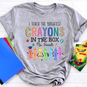 May include: Gray t-shirt with the text "I teach the brightest crayons in the box Ms. Sarah" and a row of colorful crayons with faces.
