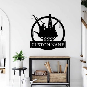 Könnte beinhalten: Schwarze Metallsilhouette einer Ölplattform mit einem Kreis darum und dem Text "CUSTOM NAME" darunter. Die Silhouette ist an einer weißen Wand montiert.