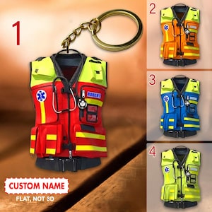 Può includere: Un portachiavi con un designo piatto in 2D di un giubbotto di sicurezza rosso e giallo con uno stetoscopio, un simbolo di stella della vita e il nome "ROBERT". Il portachiavi è disponibile in quattro colori diversi: rosso, arancione, blu e giallo.