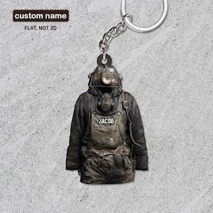 Könnte beinhalten: Ein schwarzer und grauer Schlüsselanhänger mit einem Bergmann, der einen Helm und eine Gasmaske trägt. Der Schlüsselanhänger trägt den Namen "JACOB" auf der Vorderseite.