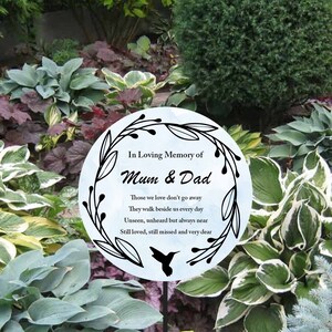 Könnte beinhalten: Ein weißer Gartenpflock mit einem schwarzen Kolibri und einem blauen Aquarellhintergrund. Der Pflock hat einen schwarzen Kranz mit Blättern und den Text "In liebevoller Erinnerung an Mama und Papa" sowie ein Gedicht über Verstorbene.