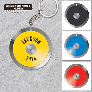 Puede incluir: Un llavero personalizado con un disco amarillo con el nombre "JACKSON" y el año "2024" en texto negro. El llavero tiene un borde de metal plateado y un pequeño centro de metal. Se muestran discos adicionales en negro, azul y rojo.