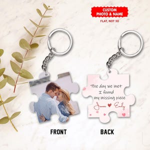 Puede incluir: Un llavero personalizado con forma de pieza de rompecabezas. La parte delantera muestra una foto de una pareja, mientras que la trasera tiene el texto "The day we met I found my missing piece" con nombres y corazones. El llavero es plateado.