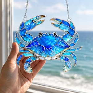 May include: A stained glass suncatcher shaped like a crab, featuring various shades of blue and white glass pieces held together by dark metal solder lines. The crab has two raised claws and multiple legs, with two small black beads serving as eyes.