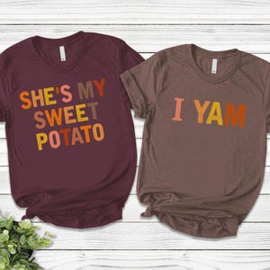 Könnte beinhalten: Zwei T-Shirts mit einem Thanksgiving-Paar-Design. Ein Shirt ist weinrot und sagt "She's my sweet potato" in Orange, Rot und Gelb. Das andere Shirt ist braun und sagt "I yam" in Orange, Rot und Gelb.
