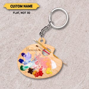 May include: A wooden artist's palette keychain with a silver keyring and chain. The palette is painted with colorful swatches of paint and includes the name "Hannah". The text "CUSTOM NAME" and "FLAT, NOT 3D" are also visible.