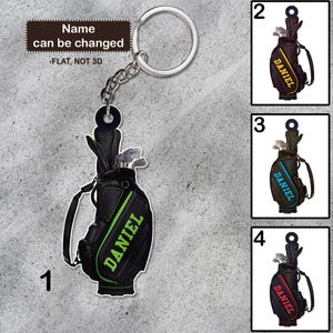 May include: A black golf bag keychain with the name "DANIEL" in green. The keychain is attached to a silver keyring. The text "Name can be changed" and "FLAT, NOT 3D" are also visible.
