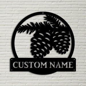 Puede incluir: Un letrero de metal negro con un diseño de piña y rama. El letrero tiene un marco circular y una placa de nombre rectangular que dice "CUSTOM NAME".