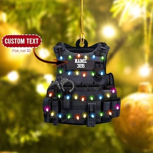 May include: A black tactical vest with a name tag that says "NAME 326" and a string of colorful Christmas lights wrapped around it. The vest is a Christmas ornament.