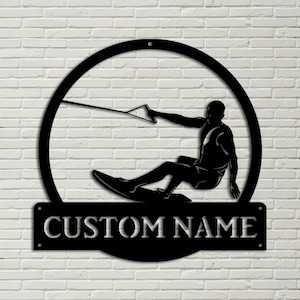 May include: Black metal silhouette of a person wakeboarding with a rope attached to a tower. The silhouette is inside a circle with a black metal sign below that says "CUSTOM NAME".