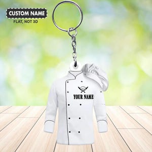 Peut inclure: Un porte-clés en forme de veste de chef blanche avec un chapeau de chef. Le porte-clés a un anneau et une chaîne argentés. La veste de chef a des boutons noirs et le texte "YOUR NAME" sous des couteaux croisés. Le texte "CUSTOM NAME" est au-dessus.