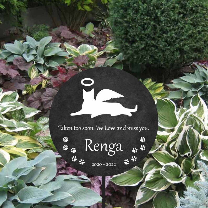 May include: Black memorial garden stake with a white silhouette of a cat with angel wings. The text reads "Taken too soon. We Love and miss you. Renga 2020 - 2022".