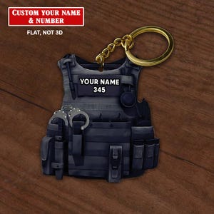 May include: A black tactical vest keychain with a gold chain. The vest has a name tag that says "YOUR NAME 345".