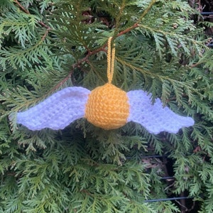 May include: A crocheted golden snitch ornament with white wings. The ornament is hanging from a tree branch.