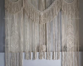 May include: A cream-colored macrame wall hanging with geometric patterns and fringe. The textile art piece features a decorative top section with a diamond-shaped design and long vertical strands. The piece is displayed against a gray wall.
