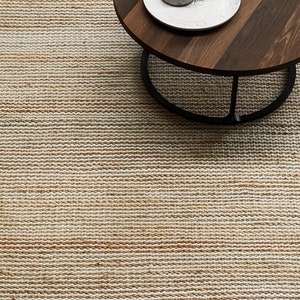 May include: A close-up of a natural fiber rug with a subtle ombre effect, transitioning from a light beige to a warm brown. The rug is woven with a textured, slightly raised pattern. A wooden coffee table with a black metal base sits on top of the rug.