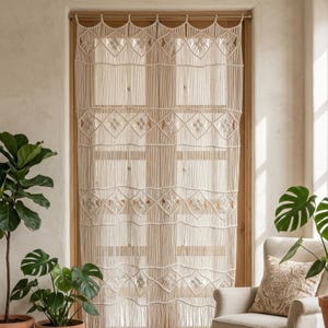May include: Cream-colored macrame window curtain. The curtain features a geometric design with diamond and floral patterns. The curtain hangs in front of a window with a wooden frame. The room has plants and a neutral color scheme.