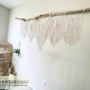 May include: A cream-coloured macrame wall hanging, suspended from a natural driftwood branch. The handcrafted piece features intricate knotting patterns and fringe, adding a bohemian touch to the neutral-toned wall. A woven shelf and hanging plant are also visible.