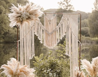May include: A white macrame wedding arch with a large white pampas grass bouquet on each side. The arch is set up outdoors in front of a lake and trees.