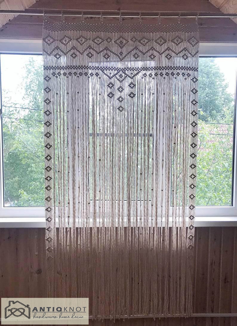 May include: A macrame curtain made of natural cotton cord hangs in a window. The curtain features a geometric pattern at the top and is made of a series of knotted cords.