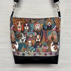 May include: A crossbody bag featuring a colorful fabric print of various dog breeds with geometric patterns. The bag has a black strap with silver metal clips, a horizontal ribbon trim with small paw prints, and a solid black base panel.
