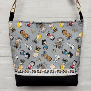 May include: A gray crossbody bag featuring a repeating pattern of cartoon characters. The bag has a black bottom and a black strap with gold-colored hardware. The word "FRIEND" is printed along the bottom edge of the bag in a repeating pattern.