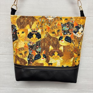 May include: A shoulder bag with a cat-themed design. The upper portion features a repeating pattern of cats in various colors, set against a gold and brown background. The bottom section is black. The bag has a black strap with gold-colored hardware.