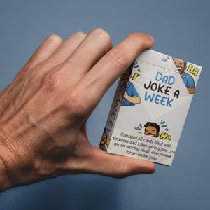 May include: A white card box labelled "DAD JOKE A WEEK" is held in a hand. The box features cartoon illustrations and the text "Contains 52 cards filled with timeless dad jokes, giving you one groan worthy laugh every week for an entire year."