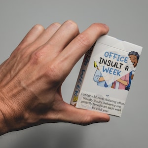 Może przedstawiać: Białe pudełko z grą karcianą zatytułowane "OFFICE INSULT A WEEK" trzymane w dłoni. Pudełko zawiera ilustracje pracowników biurowych i tekst "Zawiera 52 karty z przyjaznymi dla biura obelgami... na cały rok."